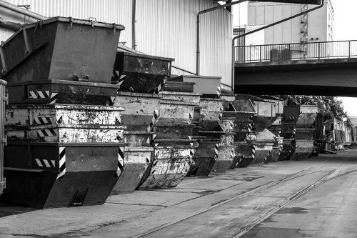 A row of large industrial skips made of metal with visible rust and weathering marks, positioned along a concrete surface in an outdoor area of a commercial or industrial site. The skips are black and grey, with some showing peeling paint and corrosion, and are partially filled or empty. They are aligned side by side, with the open tops facing upward, and are set against a backdrop of a large warehouse or factory building with metal siding, on the left side of the image. Overhead, there are pipes and a balcony or walkway extending from the building, with some shadows cast on the ground, suggesting daylight. The environment appears clean but utilitarian, characteristic of waste management or rubbish disposal facilities, with ample space for loading waste items. This scene could be part of services offered by Commercial Waste Haringey in London, supporting local businesses with rubbish removal and waste collection solutions.