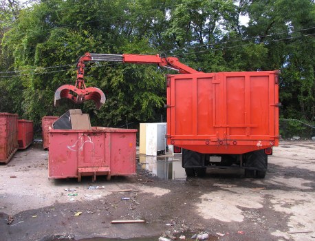 The image depicts a red waste collection truck fitted with a mechanical arm, positioned next to a large, open-top red dumpster that appears to be used for rubbish collection in an outdoor area. The truck's body and the waste container both show signs of wear and are painted in a bright red finish, contrasting with the surrounding environment. The mechanical arm is actively engaged, extending over the dumpster with a grab attachment, likely in the process of lifting or moving waste material. In the background, a green canopy of trees adds a natural backdrop to the scene, with the ground surface consisting of dirt and scattered debris, typical of an operational waste management site. To the left, additional red bins are visible, suggesting a series of collection points, while the general environment is consistent with a municipal or commercial waste collection setting, such as in Haringey. The image emphasizes a practical rubbish removal operation conducted by a waste management service, represented here by Commercial Waste Haringey, within a typical urban or semi-urban outdoor space dedicated to rubbish disposal and collection activities.