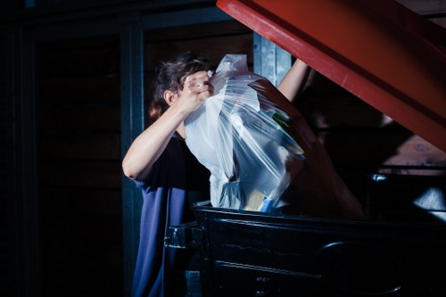 A person is seen loading clear plastic rubbish bags filled with miscellaneous waste into a large black industrial waste bin on a paved outdoor area. The individual, whose face is partially obscured, is wearing a dark short-sleeved shirt and is standing next to the bin, which is situated against a backdrop of wooden and metal fencing. The waste bags appear to contain various household or commercial refuse, with some items visibly protruding through the plastic. The surrounding environment suggests an outdoor space, possibly a back garden or driveway, with natural lighting casting shadows on the scene. This image is relevant to rubbish removal services provided by Commercial Waste Haringey, based in the Haringey area, emphasizing the process of collecting and disposing of mixed waste materials.