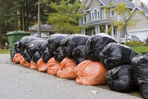 A row of black and orange plastic rubbish bags stacked along the edge of a residential street on a paved pavement. The black bags, appearing to be made of thick, durable plastic, are gathered tightly and tied at the top, with their glossy surfaces reflecting ambient light. The orange bags, likely used for waste collection or disposal, are situated underneath some of the black bags, partially visible, and have a slightly softer, matte finish. The bags are aligned in a single file, leaning against a curb, with some appearing slightly bulging from contained rubbish. In the background, a green wheelie bin is positioned on the pavement, beside a driveway leading to a modern, painted house surrounded by greenery, trees, and other parked cars, hinting at a suburban environment possibly within Haringey. The scene suggests a typical waste collection or cleanup activity carried out by a rubbish removal service like Commercial Waste Haringey, with the bags placed neatly along the street for collection. Natural daylight illuminates the scene, highlighting the textures and colors of the waste bags and surrounding environment.