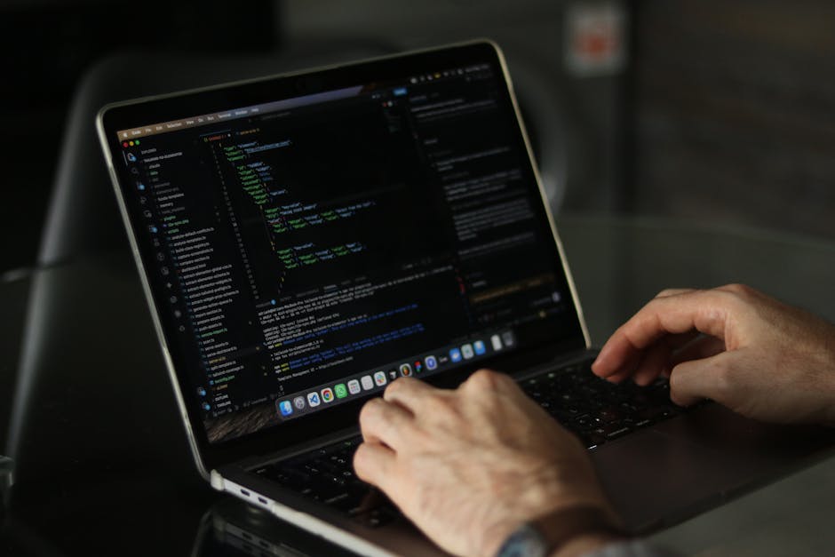 A close-up view of a person's hand operating a black computer keyboard on a white desk, with a large computer monitor displaying lines of code or terminal output in the background. The workspace inclu