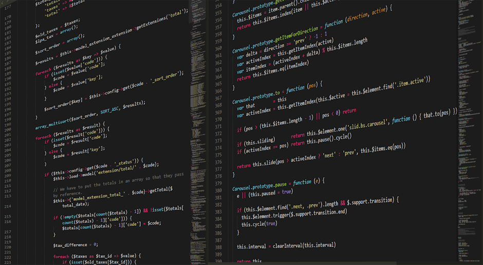 A close-up view of a computer workstation featuring a large monitor displaying lines of colourful computer code in shades of white, yellow, and teal on a dark background. The monitor is positioned on 