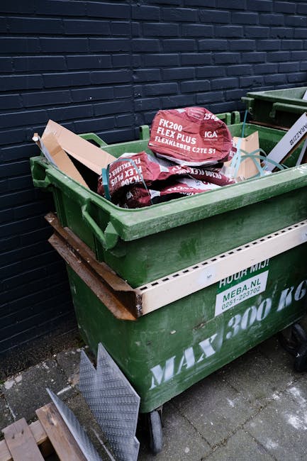 A green wheeled waste container positioned outdoors on a concrete surface beside a dark brick wall. The container is filled with red and black bags of building materials or supplies, with some bags la
