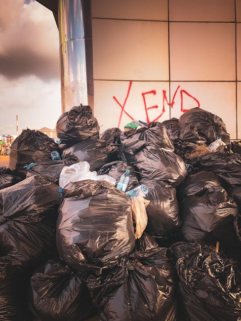 A green wheeled waste container positioned outdoors on a concrete surface beside a dark brick wall. The container is filled with red and black bags of building materials or supplies, with some bags la