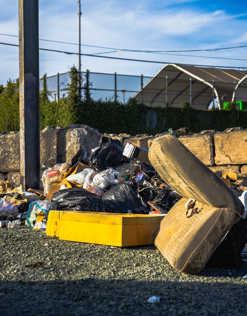Three large black rubbish bags made of plastic or thick waste material are placed on the edge of a paved street or sidewalk, leaning against a black metal fence with vertical bars. The bags appear to 